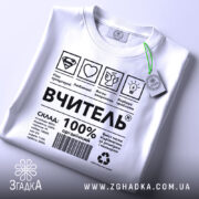 Футболка вчитель енергія, біла, складена на столі, фон світло-сірий, принт чорного кольору з символами та текстом, на футболці є етикетка з зеленим шнурком.