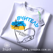 Футболка вчитель найкращого класу білого кольору, складена на поверхні, фон світло-сірий, принт з написом 'ВЧИТЕЛЬ' та малюнком серця з олівцем і квітами, на футболці є етикетка з зеленим шнурком.