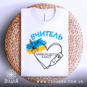 Футболка вчитель найкращого класу білого кольору, складена на плетеній кошику, фон світло-сірий, принт з написом 'ВЧИТЕЛЬ' та малюнком серця з олівцем і квітами, видно етикетку на комірці.