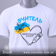 Футболка вчитель найкращого класу білого кольору, складена на поверхні, фон світло-сірий, принт з написом 'ВЧИТЕЛЬ' та малюнком серця з олівцем і квітами, на футболці є етикетка з зеленим шнурком.