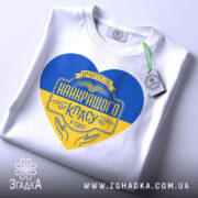 Футболка вчитель найкращого класу білого кольору, зображена зверху, на ній підвішена етикетка, фон світло-сірий, принт у формі серця з жовто-блакитними кольорами та написом 'Вчитель найкращого класу у світі'.