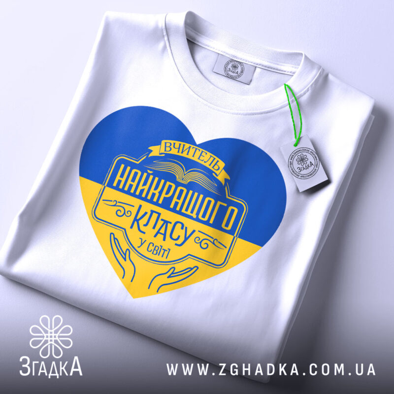 Футболка вчитель найкращого класу білого кольору, зображена зверху, на ній підвішена етикетка, фон світло-сірий, принт у формі серця з жовто-блакитними кольорами та написом 'Вчитель найкращого класу у світі'.