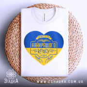 Футболка вчитель найкращого класу білого кольору, зображена зверху на плетеній кошику, фон світло-сірий, принт у формі серця з жовто-блакитними кольорами та написом 'Вчитель найкращого класу у світі'.
