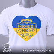 Футболка вчитель найкращого класу білого кольору, зображена зверху, на ній підвішена етикетка, фон світло-сірий, принт у формі серця з жовто-блакитними кольорами та написом 'Вчитель найкращого класу у світі'.