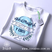 Футболка вчитель у світі, біла, зображена зверху, фон світло-сірий, принт з написом 'НАЙКРАЩИЙ ВЧИТЕЛЬ У СВІТІ' у блакитному кольорі, з етикеткою на зеленій стрічці.