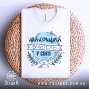 Футболка вчитель у світі, біла, зображена зверху на плетеному кошику, фон світло-сірий, принт з написом 'НАЙКРАЩИЙ ВЧИТЕЛЬ У СВІТІ' у блакитному кольорі з елементами зелені.