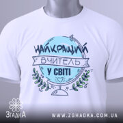 Футболка вчитель у світі, біла, зображена зверху, фон світло-сірий, принт з написом 'НАЙКРАЩИЙ ВЧИТЕЛЬ У СВІТІ' у блакитному кольорі, з етикеткою на зеленій стрічці.