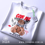 Футболка ведмедик хуліган білого кольору, складена на поверхні, з принтом ведмедика в космічному шоломі, на фоні світло-сірого кольору, деталі принта включають червоні та чорні елементи з написом 'GIVE ME SPACE EAT', з прикріпленою етикеткою.