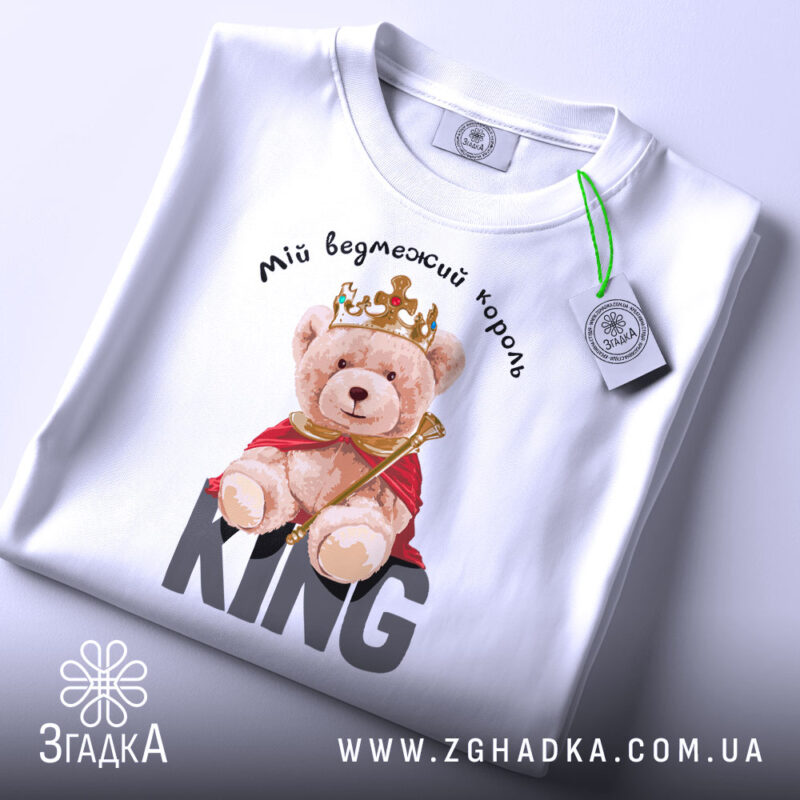 Футболка ведмежий король, білого кольору, складена на поверхні, фон світло-сірий, принт з ведмедиком у короні та червоному плащі, текст 'KING' великими літерами під ведмедиком, на футболці є етикетка.