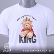 Футболка ведмежий король, білого кольору, складена на поверхні, фон світло-сірий, принт з ведмедиком у короні та червоному плащі, текст 'KING' великими літерами під ведмедиком, на футболці є етикетка.