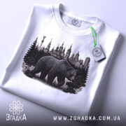 Футболка ведмідь Київ, біла, складена на поверхні, фон світло-сірий, принт з ведмедем та архітектурою Києва, з етикеткою зеленого кольору.