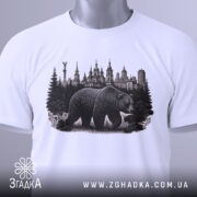 Футболка ведмідь Київ, біла, складена на поверхні, фон світло-сірий, принт з ведмедем та архітектурою Києва, з етикеткою зеленого кольору.