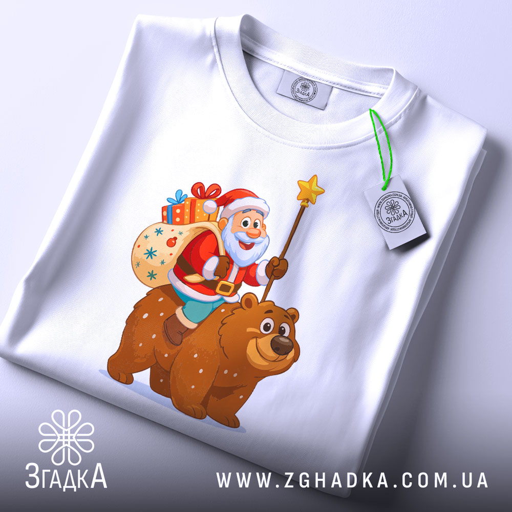 Футболка ведмідь новий рік, біла, з верхнього ракурсу, складена на поверхні, принт з дідом морозом на ведмеді, з мішком подарунків, на етикетці зелена стрічка, фон світлий.