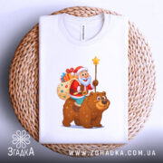 Футболка ведмідь новий рік, біла, з верхнього ракурсу, складена на плетеній кошику, принт з дідом морозом на ведмеді, фон світлий, текстильна етикетка видима.