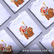 Футболка ведмідь новий рік, біла, з фронтального ракурсу, на манекені, принт з дідом морозом, який тримає зірку, та ведмедем, що несе мішок з подарунками, фон нейтральний сірий.