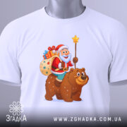 Футболка ведмідь новий рік, біла, з верхнього ракурсу, складена на поверхні, принт з дідом морозом на ведмеді, з мішком подарунків, на етикетці зелена стрічка, фон світлий.