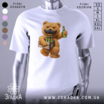 Футболка Ведмідь Пиво, біла футболка на манекені, з принтом ведмедя з вусами, який тримає ящик з пляшками пива, фон світло-сірий, деталі принта яскраві та чіткі.