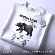 Футболка ведмідь схема, біла, розкладена на поверхні, фон світло-сірий, принт з чорними деталями, на футболці є етикетка з зеленим шнурком.