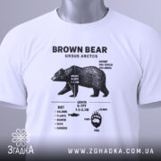 Футболка ведмідь схема, біла, розкладена на поверхні, фон світло-сірий, принт з чорними деталями, на футболці є етикетка з зеленим шнурком.