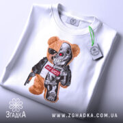 Футболка ведмідь термінатор, біла, складена на столі, фон світло-сірий, принт з ведмедем у сонцезахисних окулярах та механічною частиною, напис "НУ ЩО?" на червоному фоні, з етикеткою на зеленій стрічці.