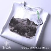 Футболка ведмідь у горах, біла, складена на поверхні, фон світлий, принт з ведмедем на фоні гір і лісу, на футболці є етикетка з зеленим шнурком.