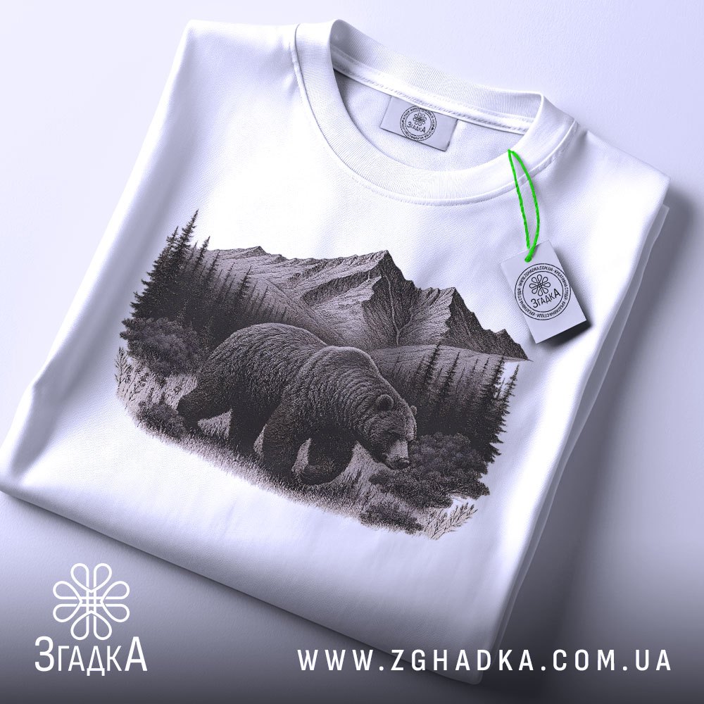Футболка ведмідь у горах, біла, складена на поверхні, фон світлий, принт з ведмедем на фоні гір і лісу, на футболці є етикетка з зеленим шнурком.