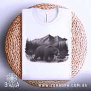 Футболка ведмідь у горах, біла, складена на плетеній кошику, фон світлий, принт з ведмедем на фоні гір і лісу, деталі принта чіткі.