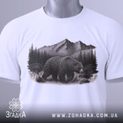 Футболка ведмідь у горах, біла, складена на поверхні, фон світлий, принт з ведмедем на фоні гір і лісу, на футболці є етикетка з зеленим шнурком.