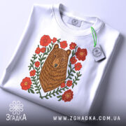 Футболка ведмідь у квітах, біла, складена на столі, фон світло-сірий, принт з ведмедем та червоними квітами, на футболці є етикетка з зеленим шнурком.