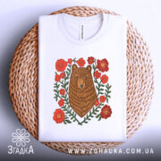 Футболка ведмідь у квітах, біла, складена на плетеному кошику, фон світлий, принт з ведмедем та червоними квітами, видно етикетку з написом.