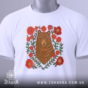 Футболка ведмідь у квітах, біла, складена на столі, фон світло-сірий, принт з ведмедем та червоними квітами, на футболці є етикетка з зеленим шнурком.