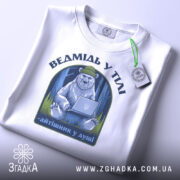 Футболка ведмідь у тілі, біла, зображена на столі, фон світло-сірий, принт з ведмедем у зеленій шапці, що сидить за ноутбуком, текст під принтом - 'айтішник у душі', на футболці є етикетка з зеленим шнурком.