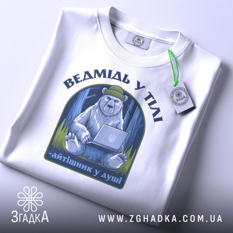 Футболка ведмідь у тілі, біла, зображена на столі, фон світло-сірий, принт з ведмедем у зеленій шапці, що сидить за ноутбуком, текст під принтом - 'айтішник у душі', на футболці є етикетка з зеленим шнурком.