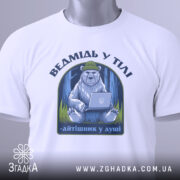 Футболка ведмідь у тілі, біла, зображена на столі, фон світло-сірий, принт з ведмедем у зеленій шапці, що сидить за ноутбуком, текст під принтом - 'айтішник у душі', на футболці є етикетка з зеленим шнурком.