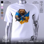 Футболка ведмідь Україна, біла, зображена на манекені, фон світло-сірий, принт ведмедя в синій та жовтій одежі, який тримає прапор України.