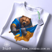 Футболка ведмідь Україна, біла, зображена на столі, фон світло-сірий, принт ведмедя в синій та жовтій одежі, з етикеткою на рукаві.