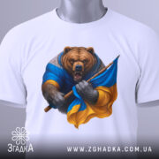 Футболка ведмідь Україна, біла, зображена на столі, фон світло-сірий, принт ведмедя в синій та жовтій одежі, з етикеткою на рукаві.
