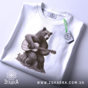 Футболка ведмідь з гітарою, біла, складена на поверхні, фон світло-сірий, принт з ведмедем, що грає на гітарі, на футболці є етикетка з логотипом.