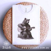 Футболка ведмідь з гітарою, біла, складена на плетеній кошику, фон світло-сірий, принт з ведмедем, що грає на гітарі, деталі принта добре проглядаються.