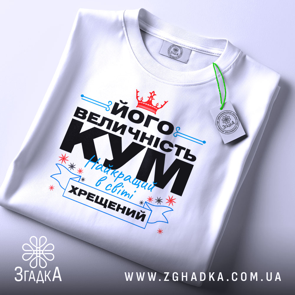 Футболка величність кум. Біла футболка, складена на поверхні, з принтом, що містить текст чорного та блакитного кольорів, а також червону корону. На футболці є етикетка з зеленим шнурком. Фон світло-сірий.