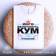 Футболка величність кум. Біла футболка, складена на плетеній основі, з принтом, що містить текст чорного та блакитного кольорів, а також червону корону. Фон натурального кольору плетеного матеріалу.