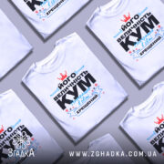 Футболка величність кум. Біла футболка на манекені, з принтом на грудях, що містить текст чорного та блакитного кольорів, а також червону корону. Фон світло-сірий, що підкреслює яскравість принта.