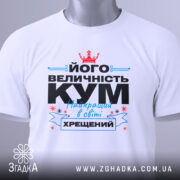 Футболка величність кум. Біла футболка, складена на поверхні, з принтом, що містить текст чорного та блакитного кольорів, а також червону корону. На футболці є етикетка з зеленим шнурком. Фон світло-сірий.
