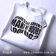 Футболка великі груди, біла, складена на столі, фон світло-сірий, принт чорний з текстом 'DOES THIS SHIRT MAKE MY BOOBS LOOK BIG?' на видному місці, з етикеткою зеленого кольору.