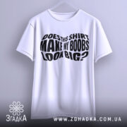 Футболка великі груди, біла, підвішена на вішалці, фон світло-сірий, принт чорний з текстом 'DOES THIS SHIRT MAKE MY BOOBS LOOK BIG?' розташований по центру.