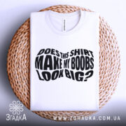 Футболка великі груди, біла, складена на плетеній кошику, фон світло-сірий, принт чорний з текстом 'DOES THIS SHIRT MAKE MY BOOBS LOOK BIG?' на видному місці.