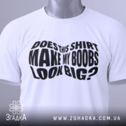 Футболка великі груди, біла, складена на столі, фон світло-сірий, принт чорний з текстом 'DOES THIS SHIRT MAKE MY BOOBS LOOK BIG?' на видному місці, з етикеткою зеленого кольору.