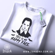 Футболка Венздей хеппі фейс білого кольору, складена на поверхні, з принтом у вигляді обличчя персонажа з косичками та написом 'THIS IS MY HAPPY FACE', фон світло-сірий, на комірці видна етикетка.