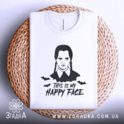 Футболка Венздей хеппі фейс білого кольору, складена на плетеній кошику, з принтом у вигляді обличчя персонажа з косичками та написом 'THIS IS MY HAPPY FACE', фон світло-сірий, деталі принта включають летючих мишей.
