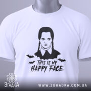 Футболка Венздей хеппі фейс білого кольору, складена на поверхні, з принтом у вигляді обличчя персонажа з косичками та написом 'THIS IS MY HAPPY FACE', фон світло-сірий, на комірці видна етикетка.